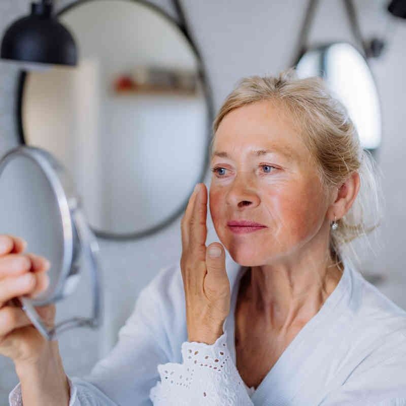mujer-madura-albura-rosa Una mujer mayor rubia con bata blanca se aplica Albura rosa crema nutritiva con vitamina A 200ml en la mejilla mientras se mira en un pequeño espejo en un baño luminoso y moderno.