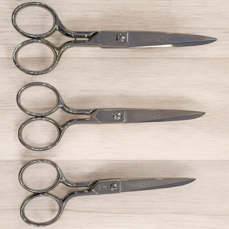 3Ctijeracosturatamainos Tres 3 Claveles tijera costura 6" (ref. 00005) están perfectamente alineados horizontalmente sobre una superficie de madera clara, los mangos a la izquierda y las hojas apuntando a la derecha.
