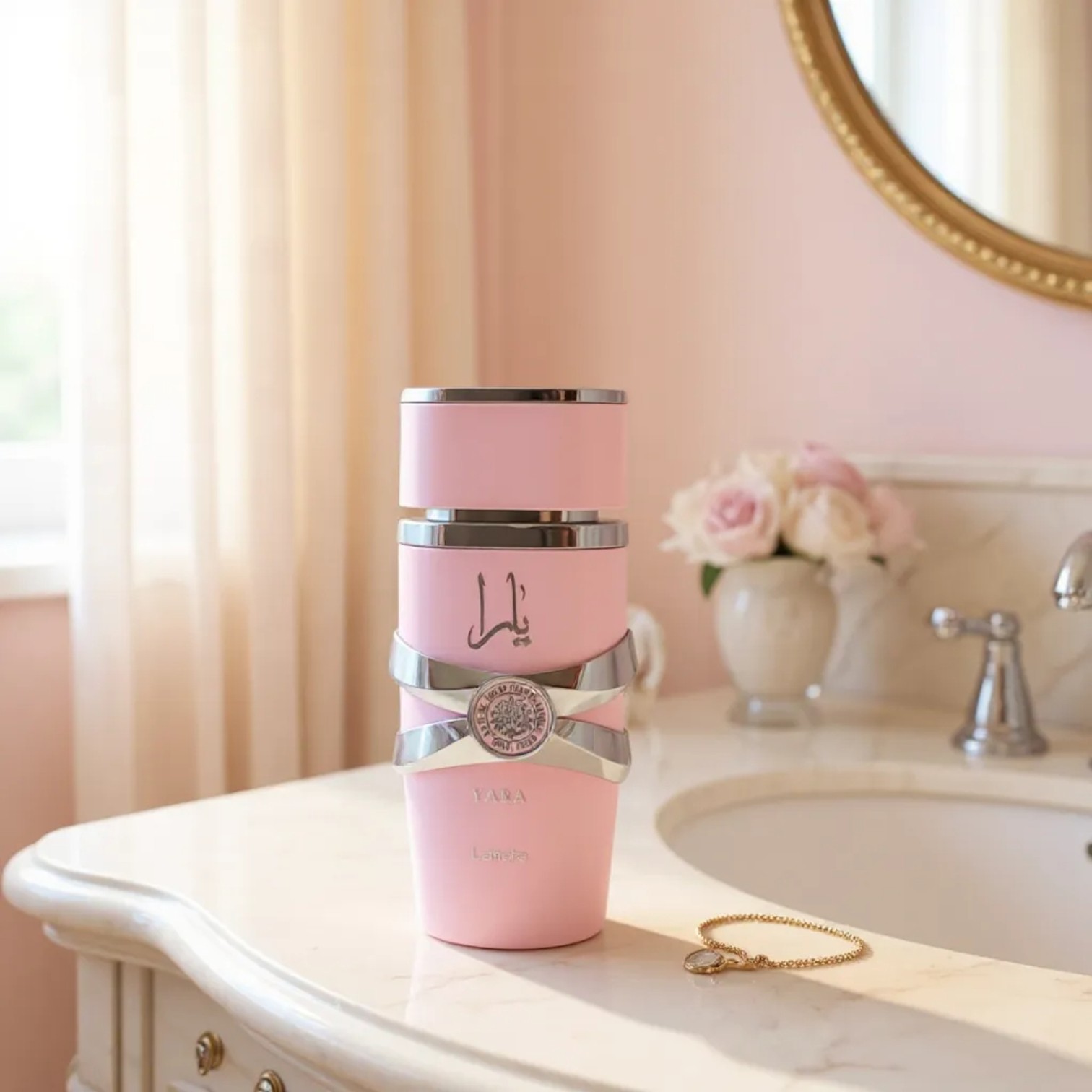 El Lattafa Yara Rosa EDP 100ml, un frasco de perfume rosa con detalles plateados, está sobre la encimera de un cuarto de baño, junto a un collar de oro y un jarrón de rosas rosas y blancas, con un espejo redondo y cortinas color crema de fondo.
