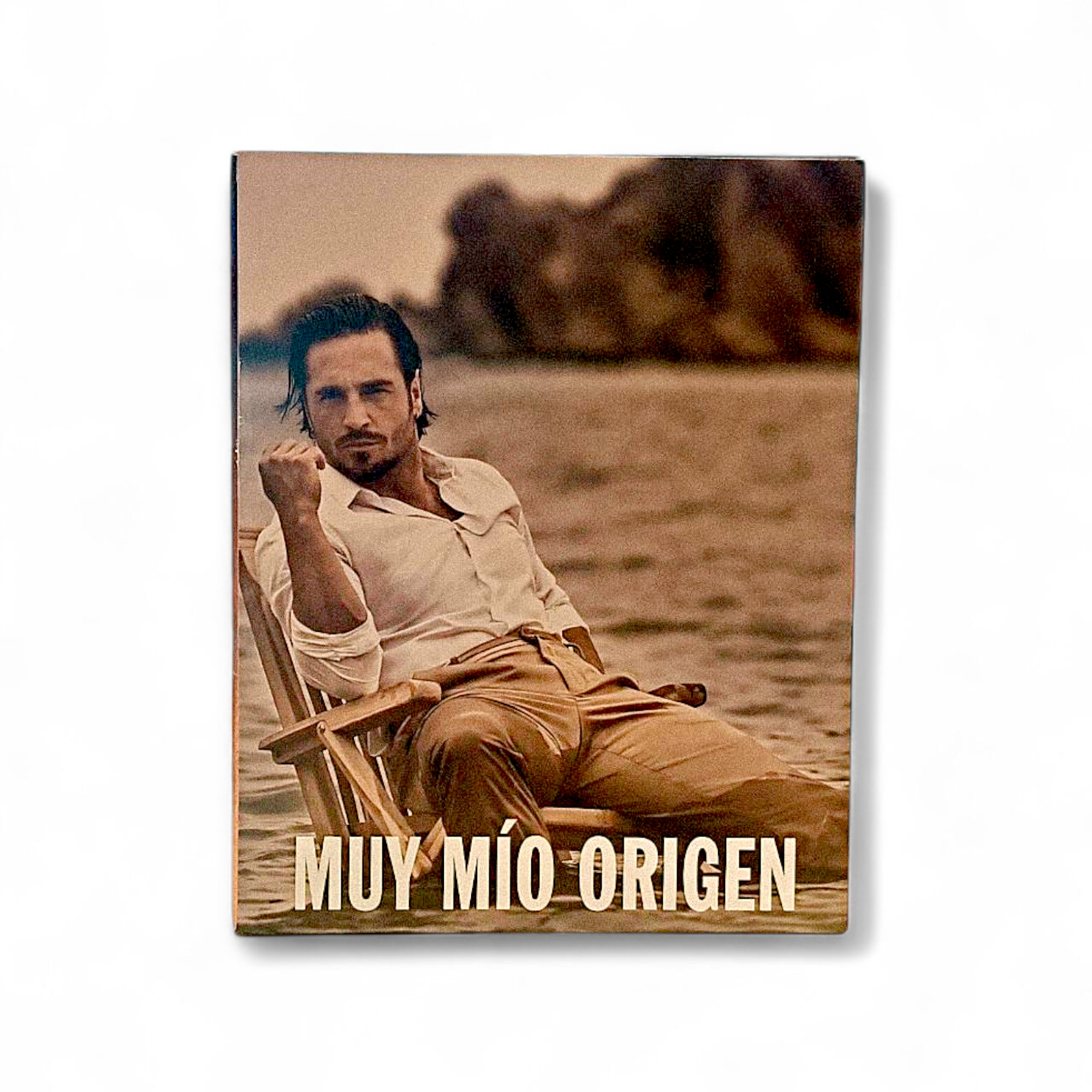La portada, en tonos sepia, muestra a un hombre con camisa blanca y pantalones color canela sentado en una silla en el agua con rocas a sus espaldas. En la parte inferior se lee "Bustamante Muy Mio Origen EDT 100ml.