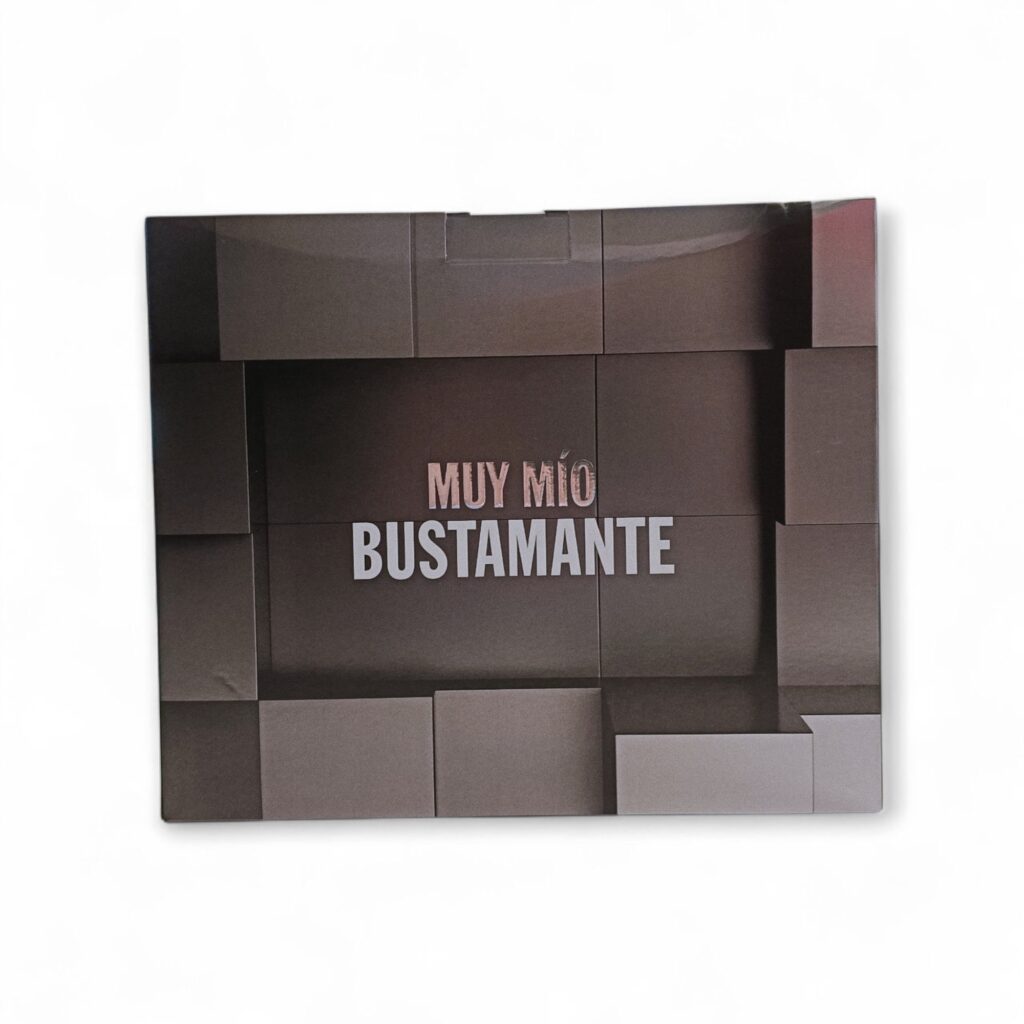 Una caja cuadrada de color marrón con motivos rectangulares en relieve y el texto «MUY MÍO BUSTAMANTE» en blanco y plata en la parte delantera contiene Bustamante Muy mio EDT 100ml + 10ml. El fondo es blanco liso. Una caja cuadrada de color marrón con motivos rectangulares en relieve y el texto "MUY MÍO BUSTAMANTE" en blanco y plata en la parte delantera contiene Bustamante Muy mio EDT 100ml + 10ml. El fondo es blanco liso.