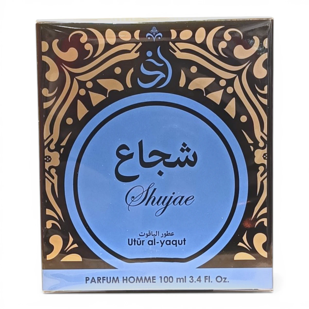 Caja de perfume con motivos ornamentales dorados y negros sobre fondo marrón, en la que figura un gran círculo azul con el texto árabe Shujae y Utür al-yaqut en inglés debajo. La caja lleva la etiqueta PARFUM HOMME 100 ml 3.4 FL. Oz.