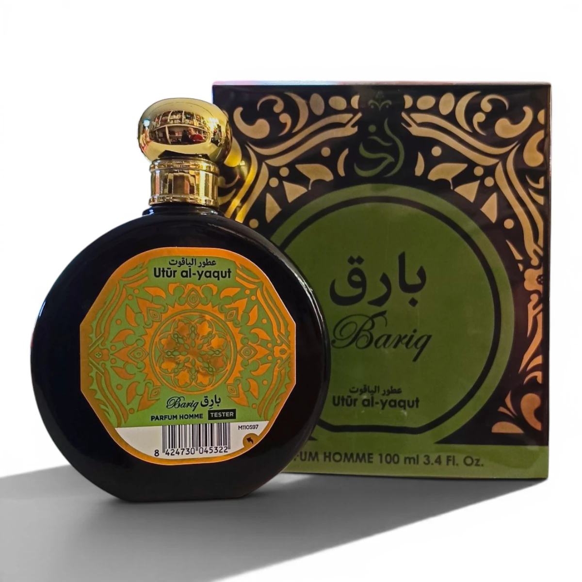 Un frasco de perfume redondo con tapón dorado y una etiqueta ornamentada en verde y naranja se encuentra delante de una caja a juego etiquetada como Bariq y Utr al-yaqat. La caja presenta motivos decorativos dorados.