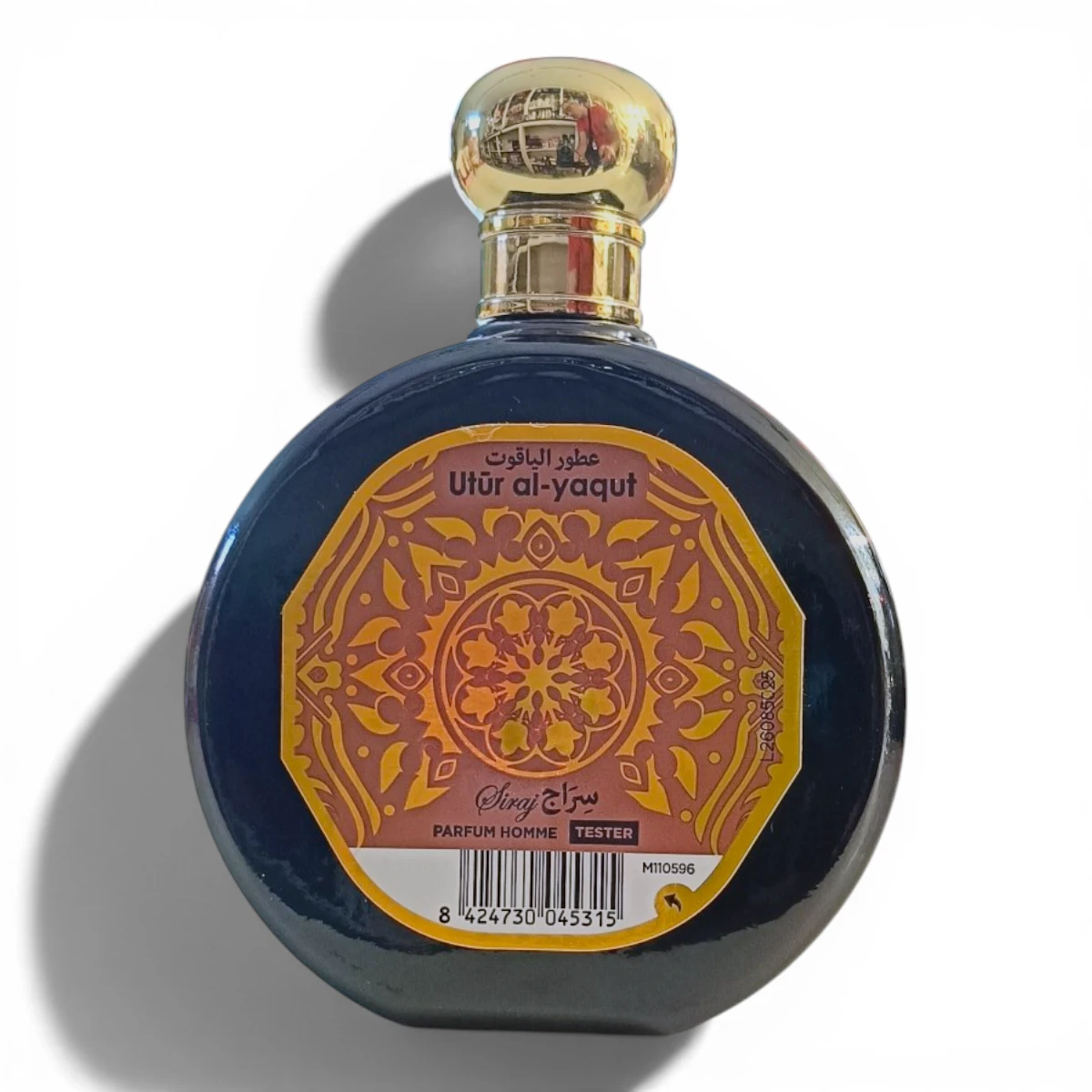 Frasco redondo de perfume con tapón dorado y etiqueta naranja y dorada ornamentada en la que se lee Utûr al-yaqut y Parfum Homme con motivos decorativos y un código de barras en la parte inferior.