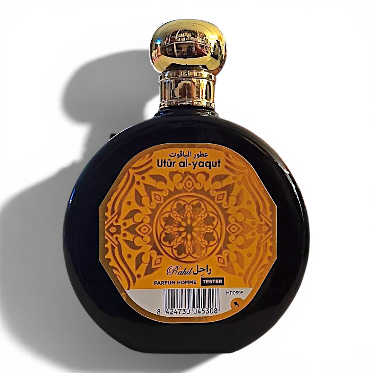Frasco redondo de perfume negro con tapón dorado y etiqueta naranja y amarilla en la que se lee Utür al-yaqut. La etiqueta presenta dibujos intrincados e incluye texto en árabe e inglés.