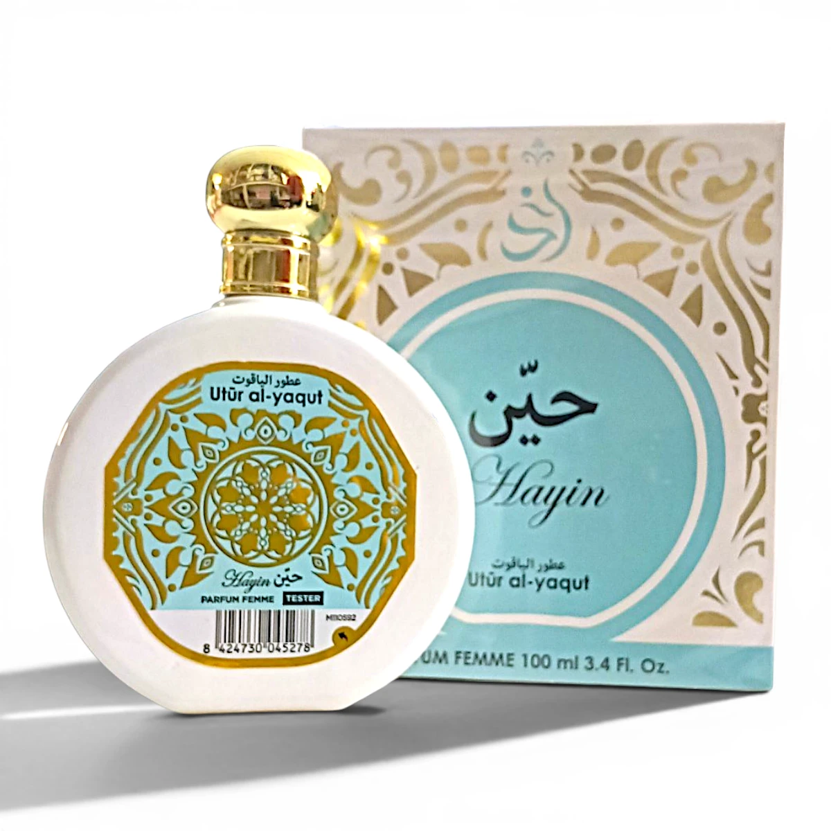 Un frasco de perfume blanco y dorado con la etiqueta Utr al-yaqut está delante de su caja ornamentada a juego, que presenta motivos decorativos en turquesa y oro y texto en árabe junto a Hayin en inglés.