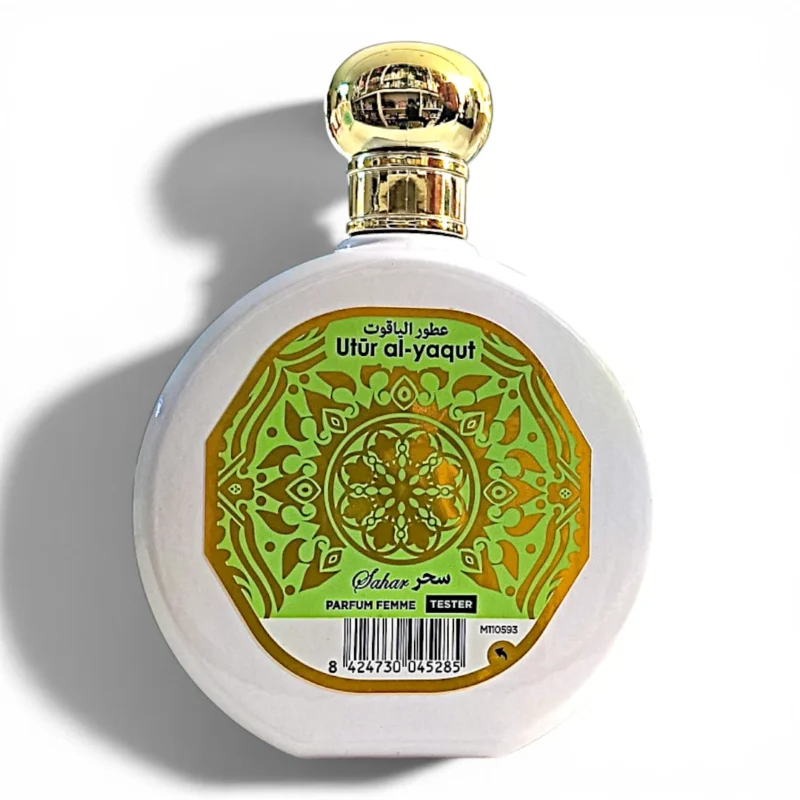 Frasco redondo de perfume blanco con tapón dorado. La etiqueta presenta diseños ornamentales verdes y dorados, texto en árabe e inglés en el que se lee Utūr al-yaqūt y Parfum Femme Tester. En la parte inferior aparece un código de barras.