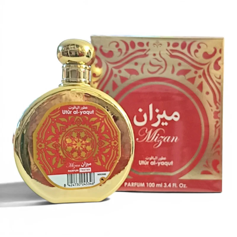 Un frasco redondo de perfume dorado con una etiqueta decorativa roja y dorada delante de una caja roja y dorada a juego con la etiqueta Mizan Utur al-yaqut. El frasco contiene 100 ml de perfume.