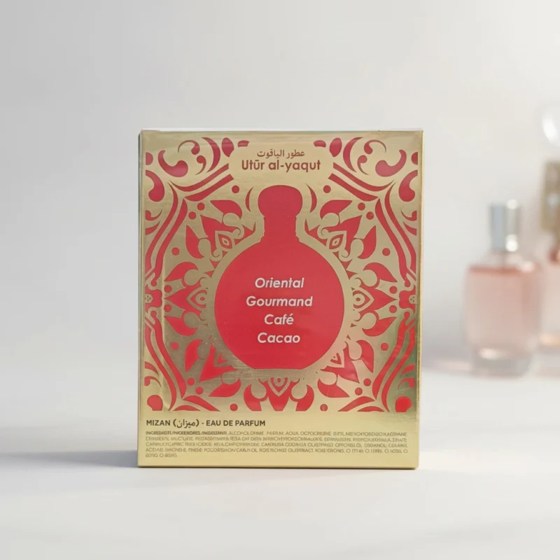 Caja de perfume dorada y roja etiquetada como Utúr al-yaqut con motivos decorativos y las notas olfativas Oriental Gourmand, Café, Cacao. En el fondo se ven frascos de perfume difuminados.