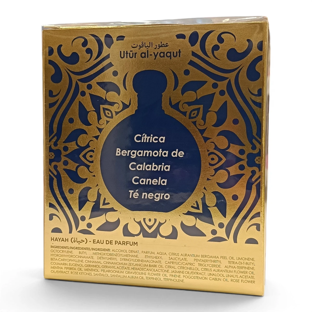 Caja de perfume dorada y azul con diseños ornamentados, texto en árabe y español y una lista de aromas: cítricos, bergamota de Calabria, canela y té negro. En la parte superior aparece la marca Utr al-yaqut.