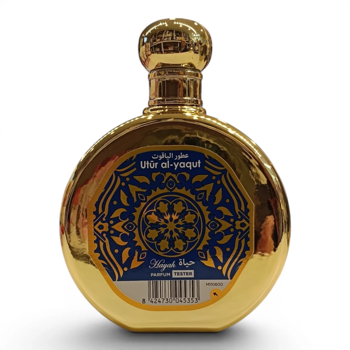 Frasco de perfume redondo, dorado, con etiqueta decorativa azul con escritura árabe y motivos ornamentales. En la etiqueta se lee Utur al-yaqut y Hayat Tester. El frasco es reflectante y se mantiene erguido.