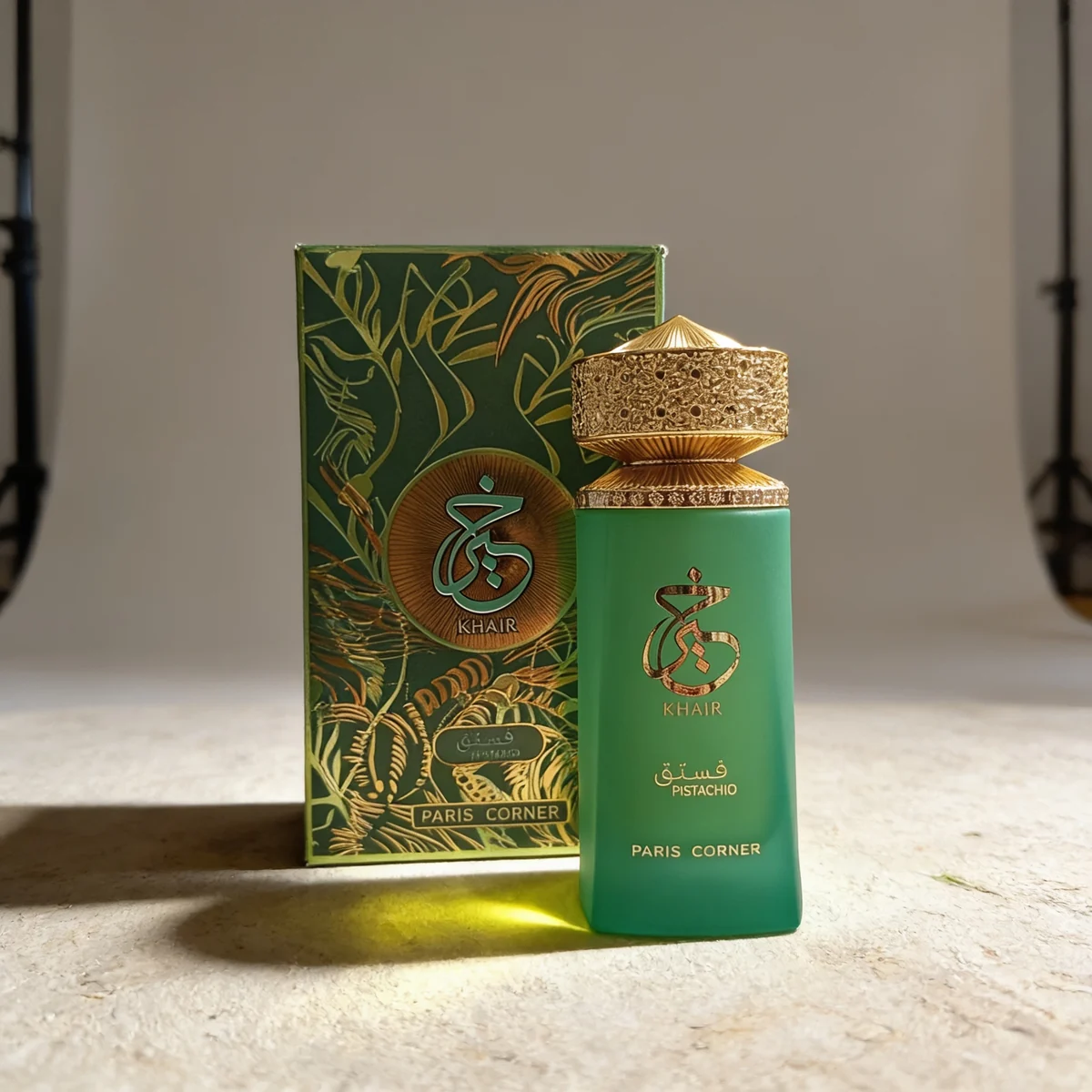 Un frasco de perfume verde y dorado con la etiqueta «Khair Pistachio Paris Corner» se encuentra frente a una caja decorativa a juego con detalles dorados y escritura árabe. La escena está suavemente iluminada en un plató de estudio.